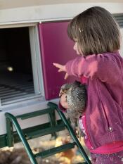 A little girl holding her pet chicken in front of a purple Eglu Cube chicken coop